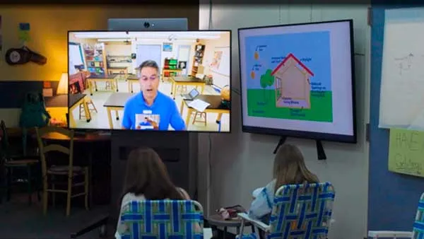 Two students sit in chairs while facing a television screen. On the screen is an adult in a classroom.