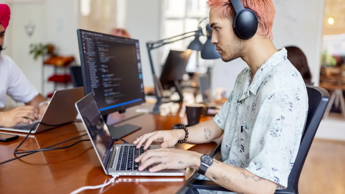 Young man wearing headphones working on computer at startup office. Young IT professional working at coworking office with people working at back.