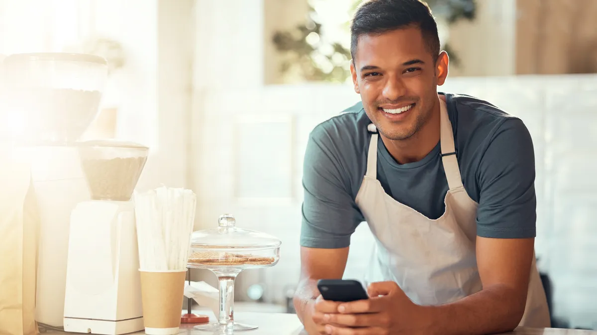 Person in blue t-shirt leaning over countertop with coffee making equipment and holding a cellphone.