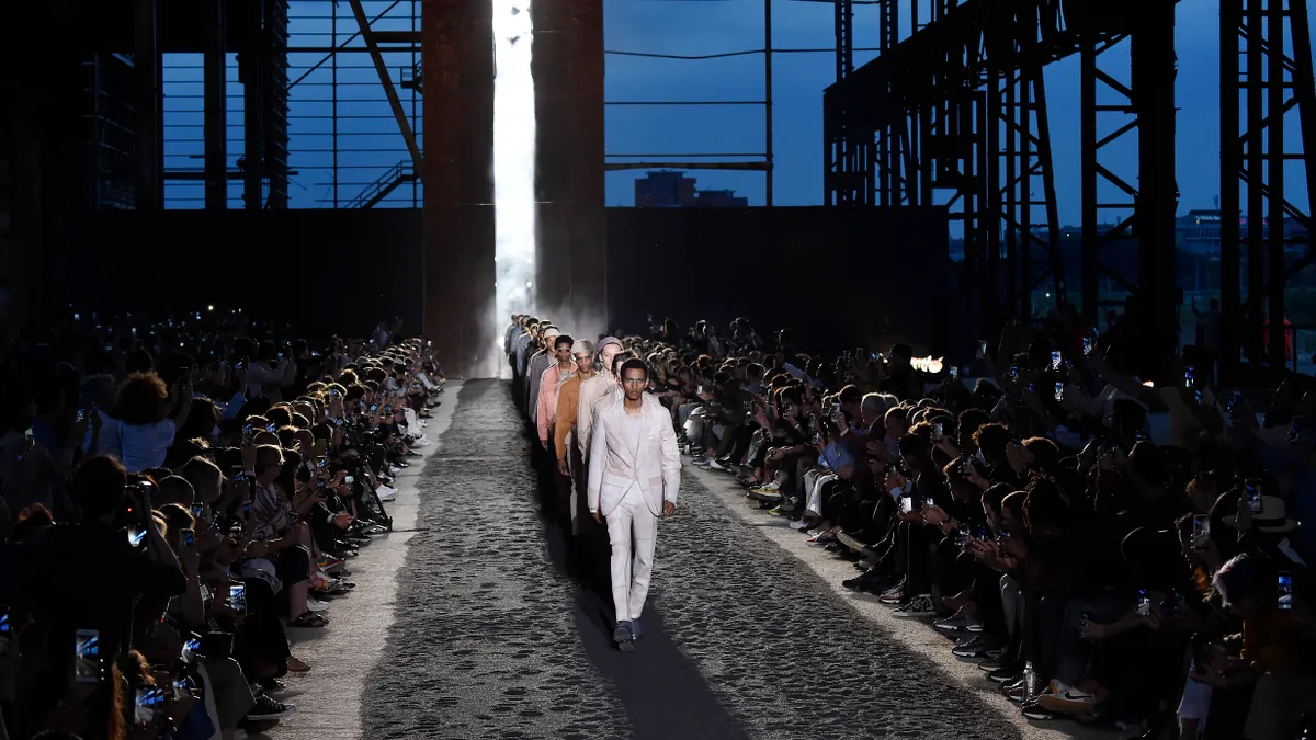 Models walk in a straight line down the runway as a crowd watches.