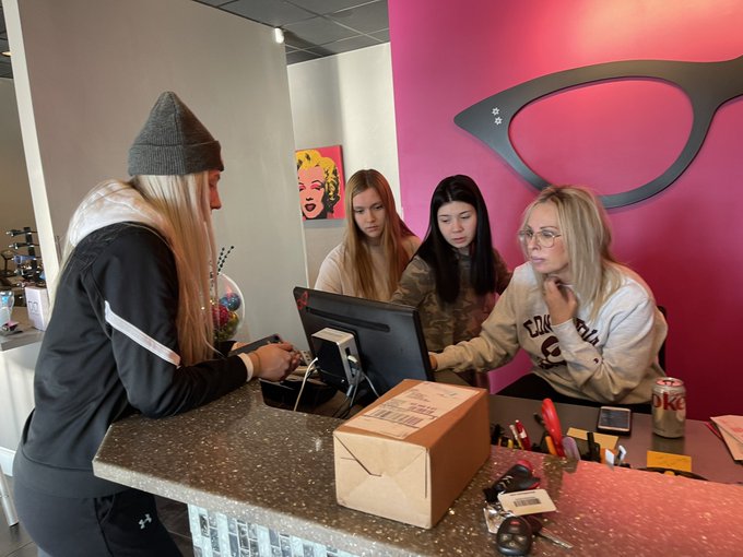 Three girl high school students and a woman optician gather around a computer at an optometry business' counter.