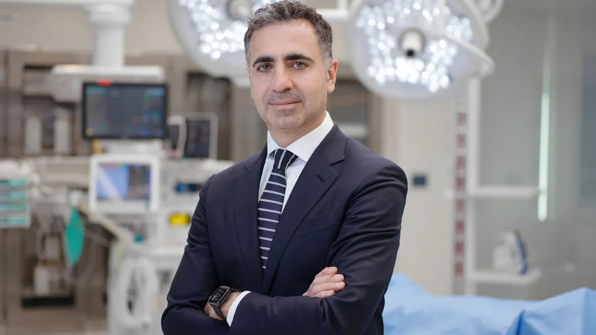 A picture of a man in a suit with his arms crossed standing in a hospital operating room.