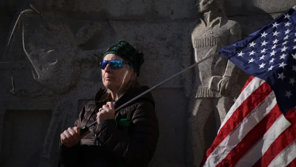 A person wearing a black coat, black hat and sunglasses holds a U.S. flag in front of a statue.
