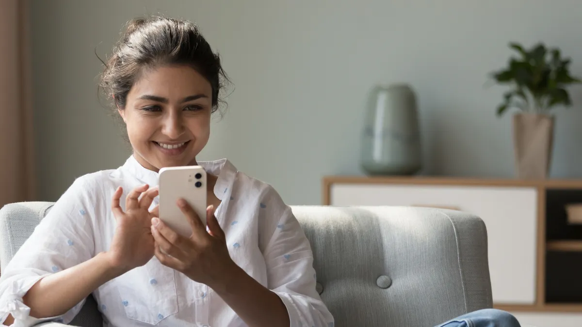 Cute Indian woman looks at cellphone screen search, web surfing information, share messages, buy goods, spend time alone at home relaxing on armchair