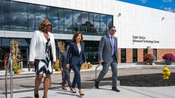 Three people in professional business attire walking, with a white and red-brick building in the background under the sun.