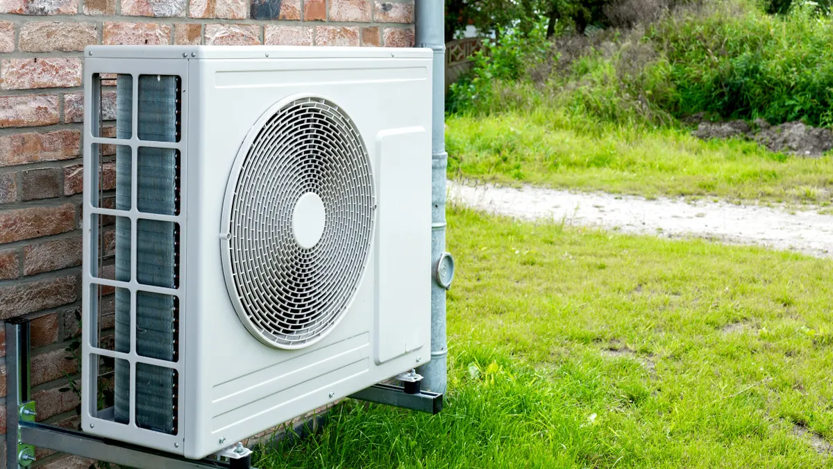 A white box with a circular fan grill sits in front of a brick house.