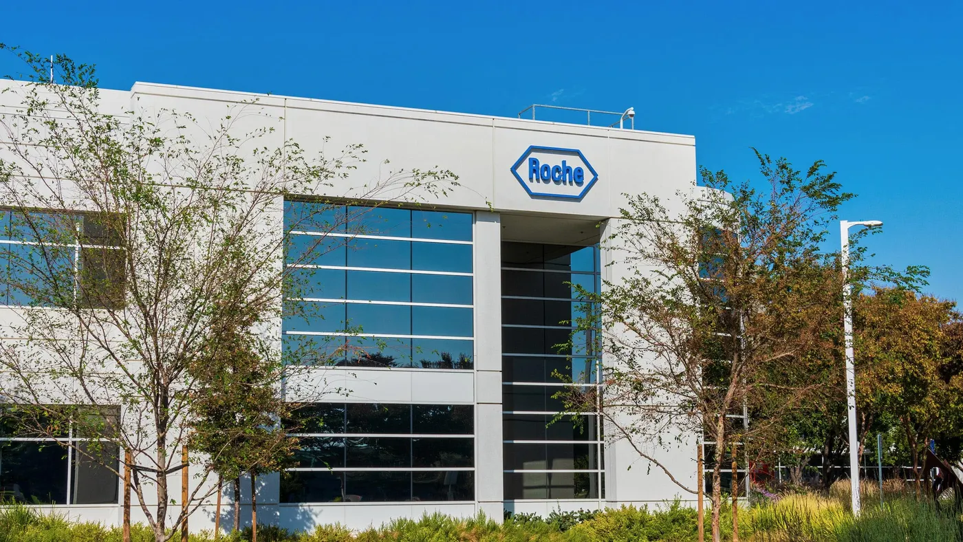 A modern office building at the Roche Diagnostics campus features a sleek glass façade with the Roche logo displayed near the entrance, and the structure is surrounded by landscaped greenery. 