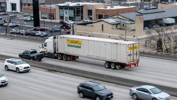 Cars and a J.B. Hunt intermodal truck drive along an interstate with buildings and a parking lot in the background.