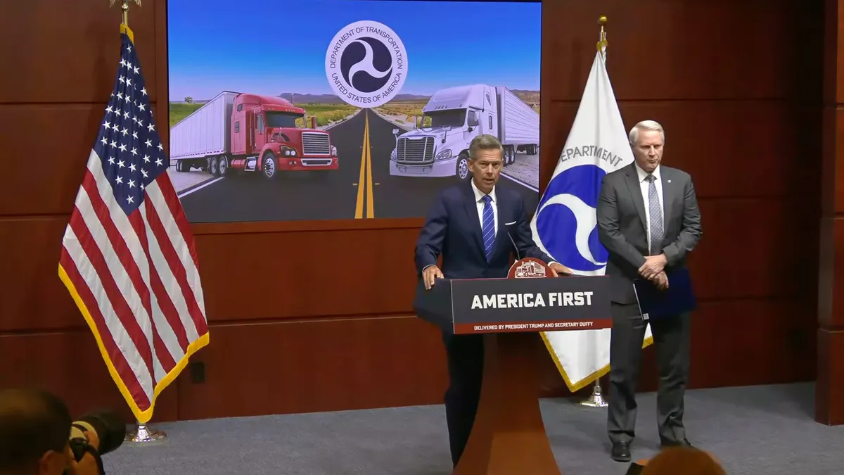 Two people stand at a podium with a sign that says, "America First." Behind them, a screen projects an image of two semi-trucks displaying the Department of Transportation logo.