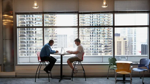 Two people on laptops sit across from each other at a table in front of a large window with Chicago skyscrapers in the background.