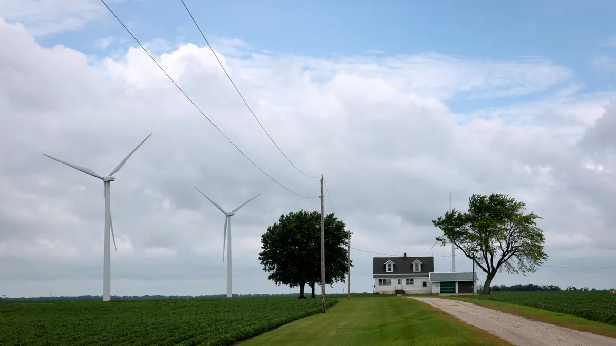 Wind turbines tower over a rural landscape in Iowa, including trees and a house.