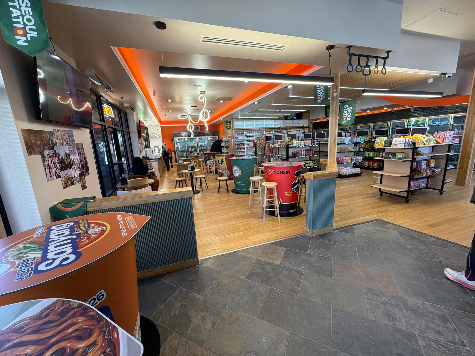 A photo looking out acorss a small convenience store and dining area. The tables are designed to look like cups of instant noodles and there are wooden stools around them. Shelves and coolers display a wide variety of goods.