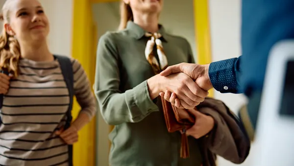 An adult is shown shaking hands with another adult while a student carrying a backpack is seen standing to the side. The hands are predominate in the photo.