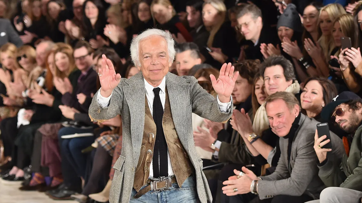 Designer Ralph Lauren, with white hair, wears a tweed jacket and a cowhide vest as he stands on the runway flanked by crowds and lifts his hands in thanks.