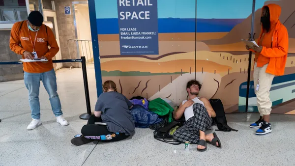 A homeless couple on the floor of a train station with two standing outreach workers wearing orange.