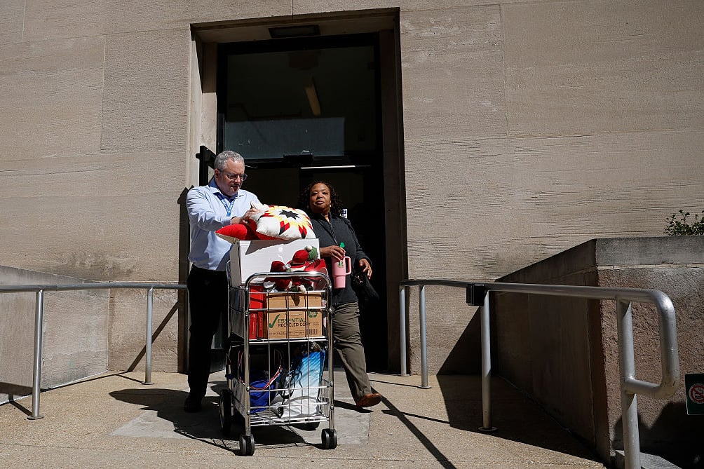 Two people wearing work attire exit a building with a cart of work items.