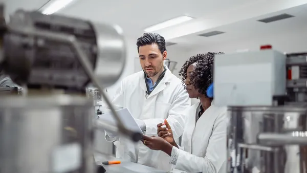Two pharmaceutical scientists in white lab coats review data in a modern laboratory.