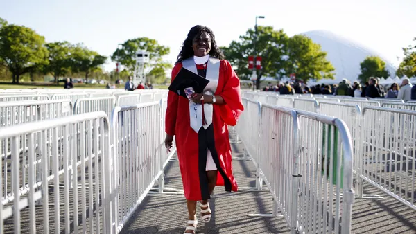 Someone wearing a graduation gown walks through crowd control lines outside.