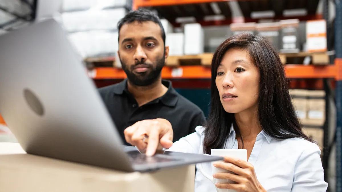 Warehouse manager using laptop to share dispatch schedule alongside a male worker. Warehouse staff looking at laptop while working.