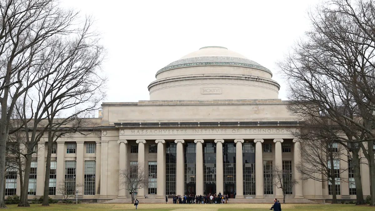 A huddle of people stand in front of a concrete building. The weather is slightly overcast, and the masonry on the building includes some Greek columns.