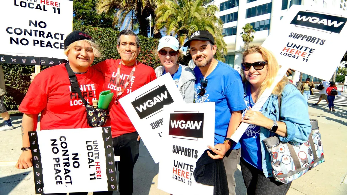 Members of Writers Guild of America West and members of Unite Here Local 11 hold picket signs on a sidewalk.