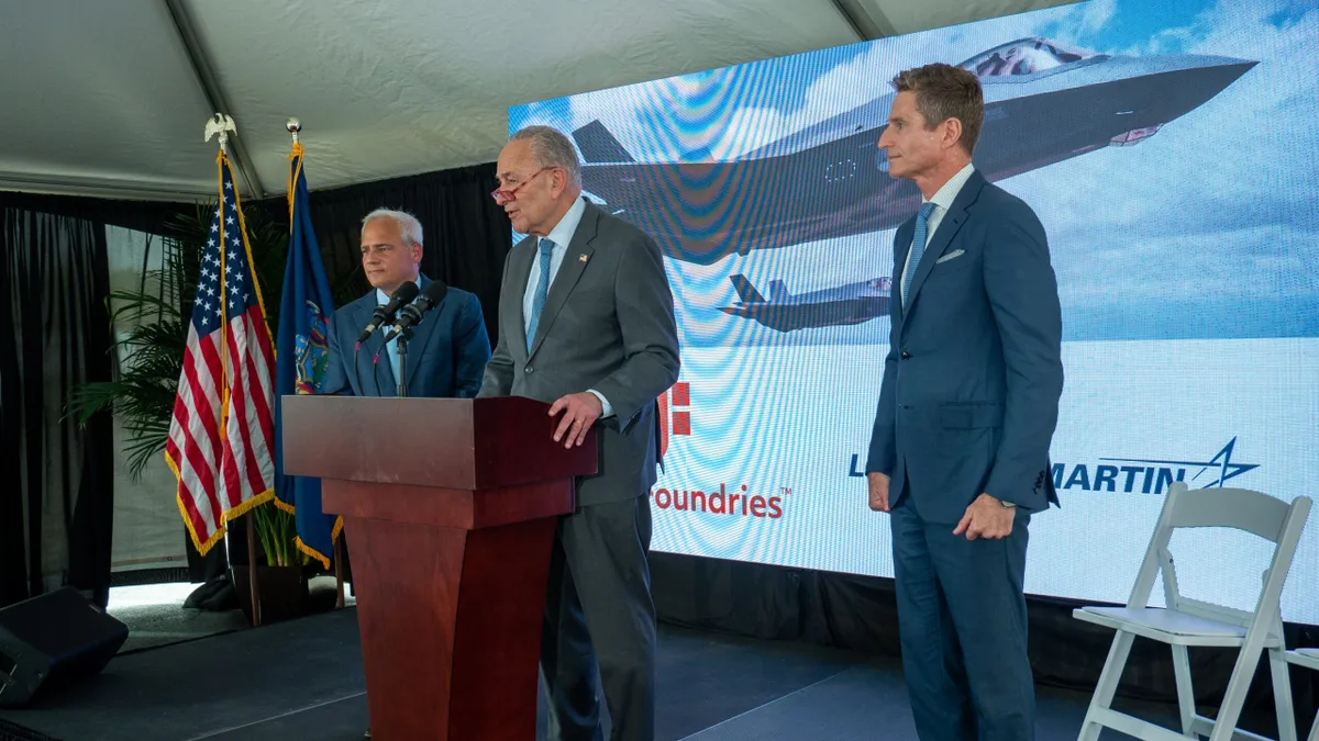 Three men standing behind a podium with a screen showing a two jets in the sky and GlobalFoundries logo on the bottom left and Lockheed Martin log on the bottom right.