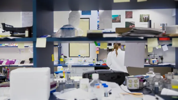 A person in a lab coat is seen through a well-lit, organized laboratory environment featuring shelves stocked with additional lab supplies and instruments. 