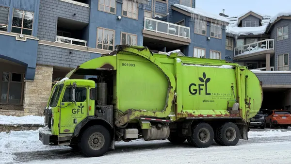 A green trash truck is parked outside a shopping complex.
