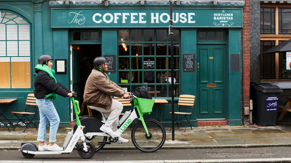 A man on a bike and a woman on a scooter ride past a coffee shop.