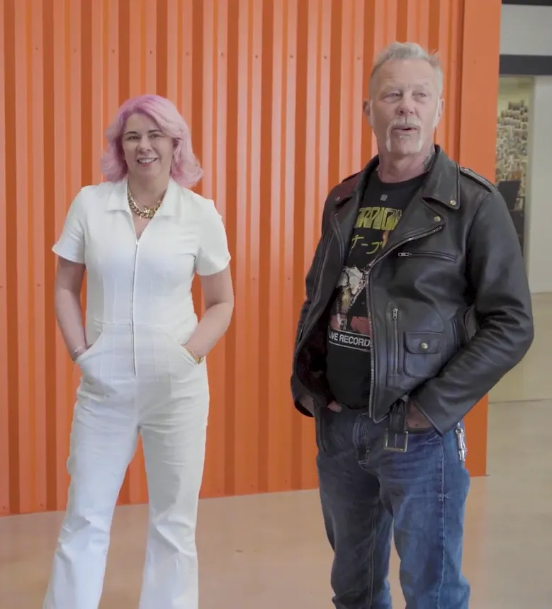 A person with pink shoulder-length hair and a white jumpsuit, standing next to a person in blue jeans, a black shirt and leather jacket.