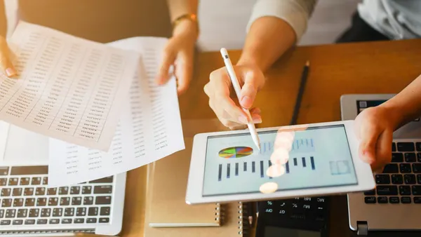 Businessman holds electronic tablet showing profit data, while businesswoman holds hard-copy report.