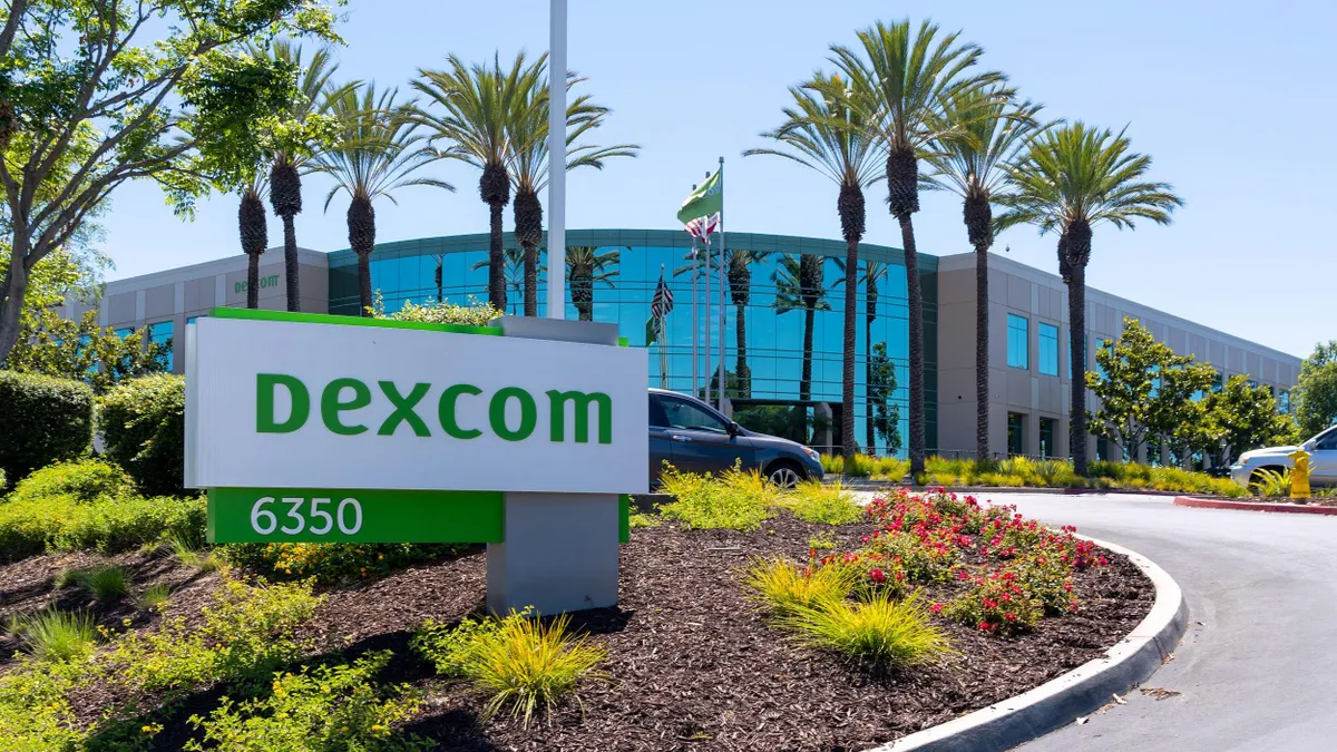 Modern office building with tall glass windows and palm trees lining the entry drive. A large white and green sign in the foreground reads “Dexcom” with the number “6350” below. Surrounding the sign are colorful landscaping features including small bushes, flowers, and a mulched bed. Cars and flags are visible in the background.