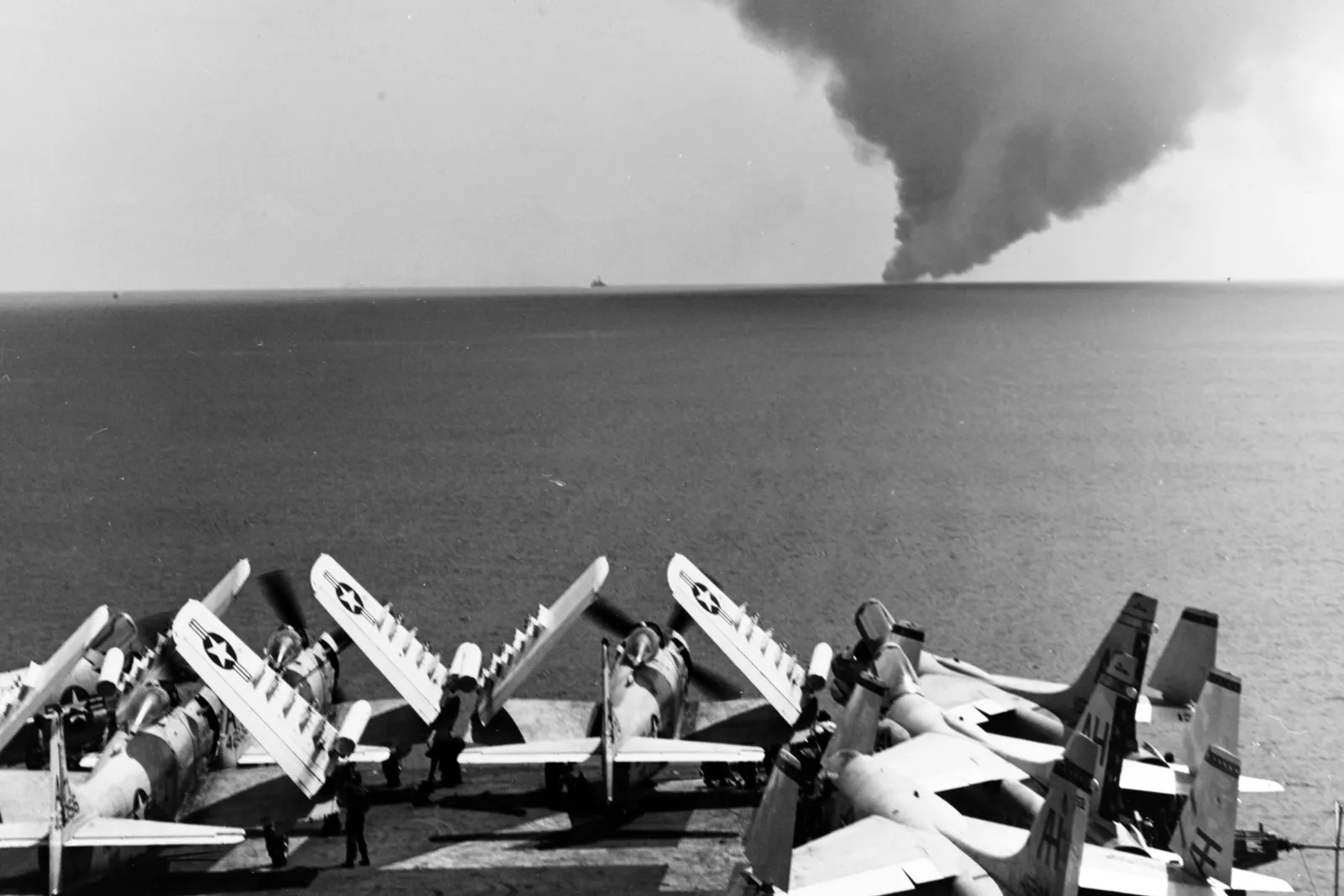 Fighter planes on a deck looking out at the sea and a large cloud of smoke.