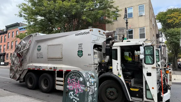 A New York City Sanitation Department rear-loading truck sits behind a graffitied mailbox on a street lined by three-story brick buildings.