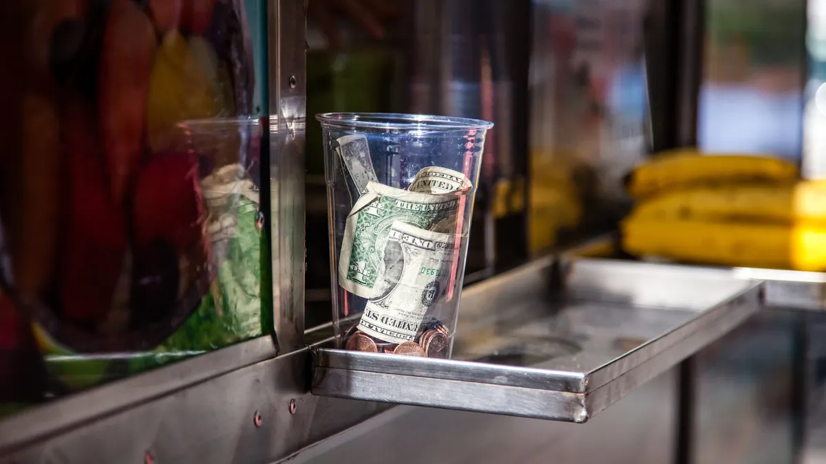 A plastic cup for tips stands on the ledge of a food cart.