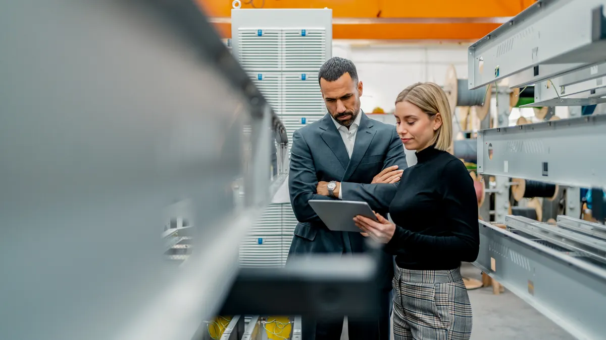 Two people stand in a modern factory, examining a tablet. One, in a suit, looks thoughtful; the other, in business casual, explains. Industrial equipment surrounds them.