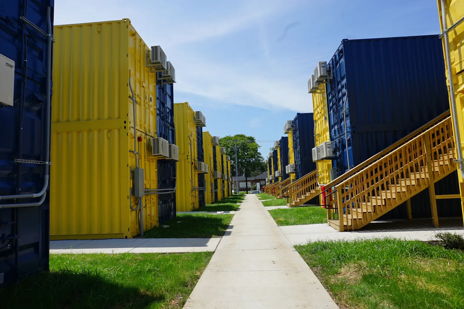 A white sidewalk with grass lawns and stacks of blue and yellow shipping container apartments on either side.