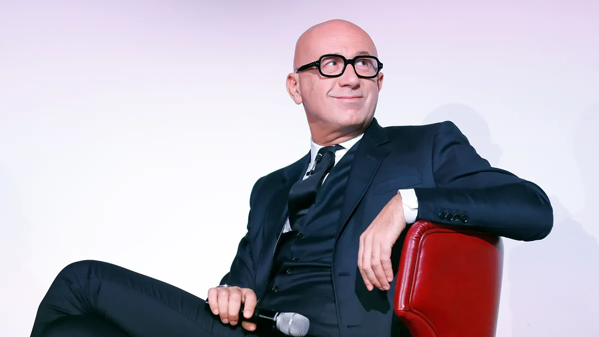 A bald man in a suit and large black glasses sits in a red chair holding a microphone and staring over his shoulder.