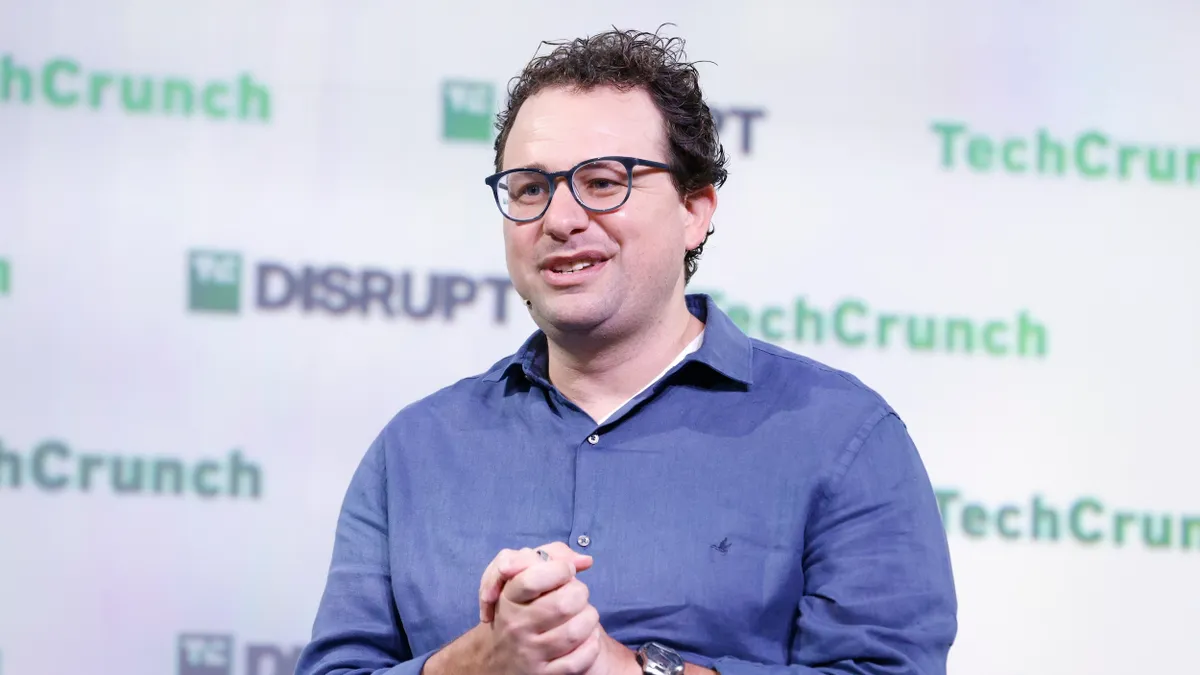 A man with curly hair and glasses and a blue button-up shirt stands on stage in front of backdrop that reads "TechCrunch" and "Disrupt."