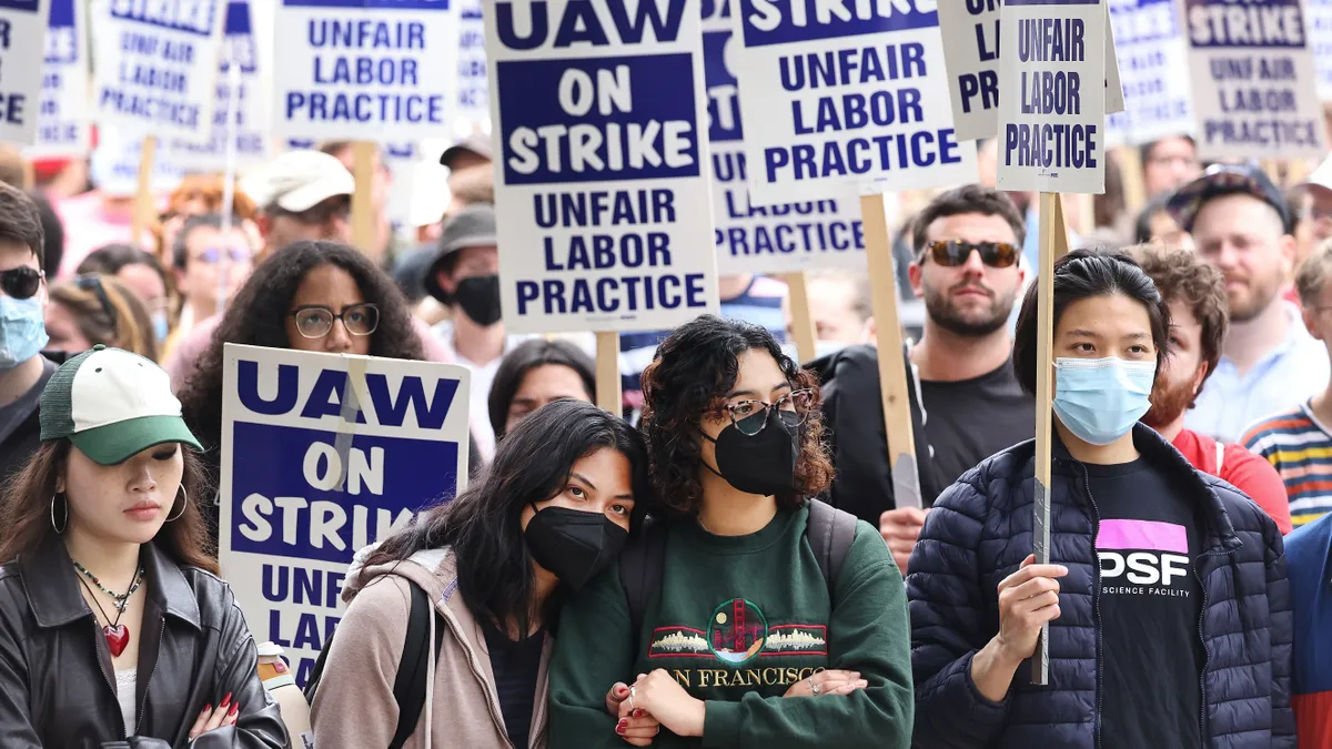 People dense crowd, some in medical masks, hold signs that say "UAW on strike, unfair labor practice."