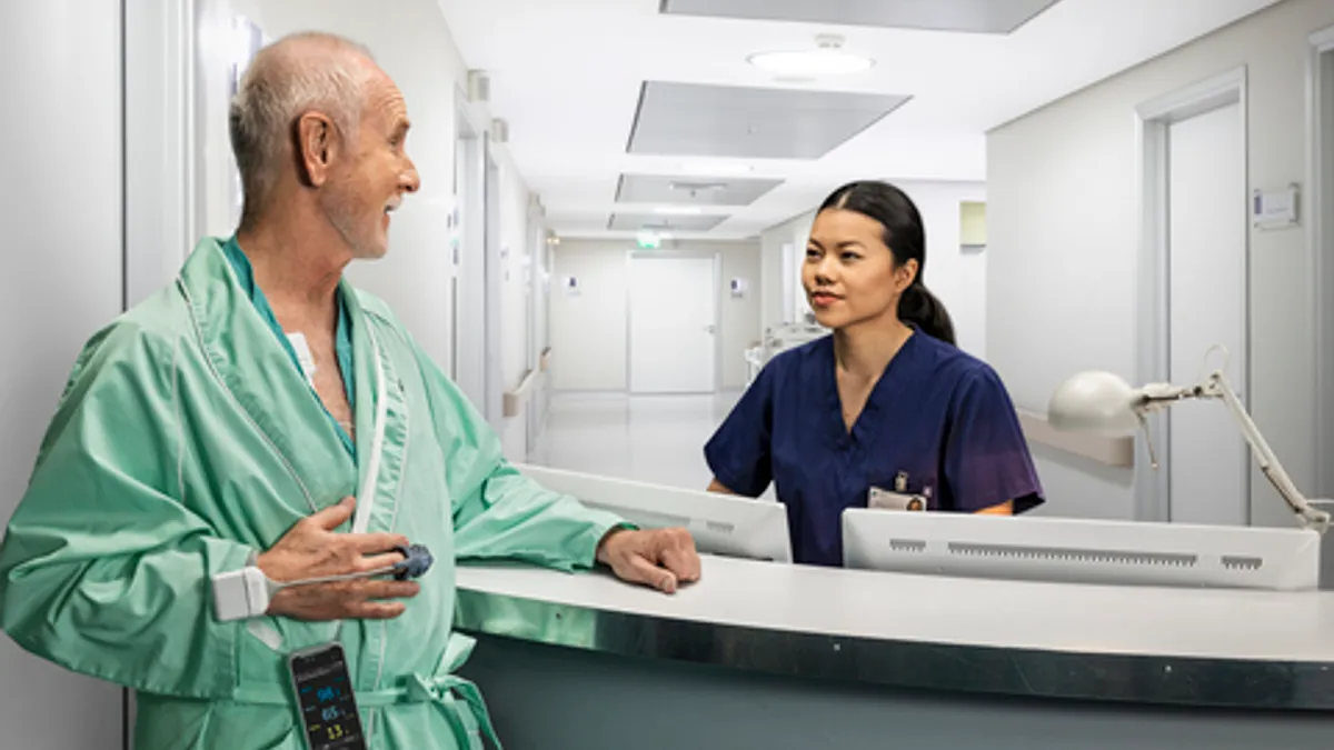 A patient wearing the GE HealthCare Portrait Mobile wireless monitoring device talks to a nurse while standing in a hospital hallway.