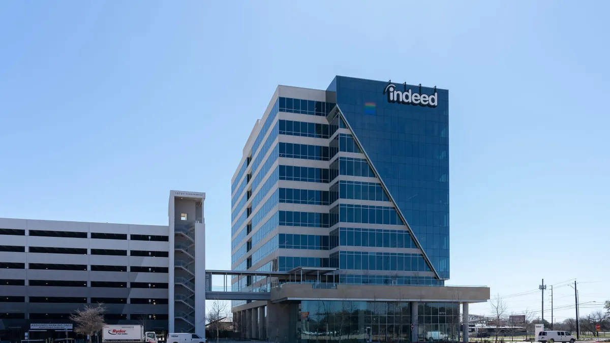 A tall glass office tower labeled “indeed” next to a multi-level parking garage and stair tower, with blue sky in the background.