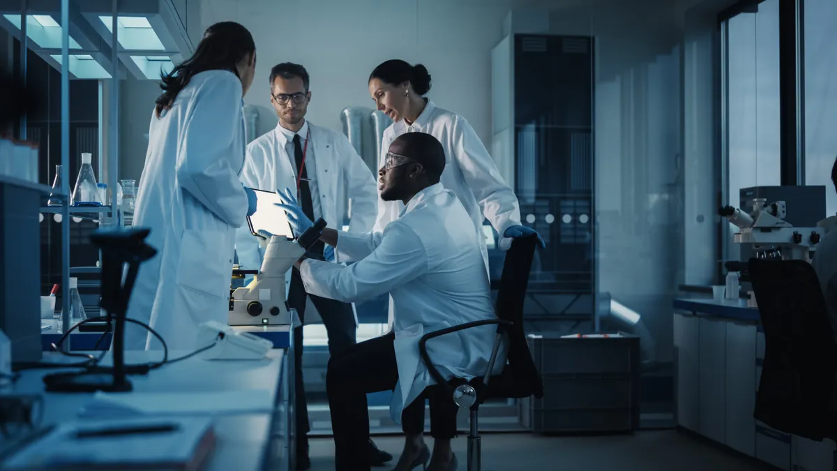 A team of four people wearing white lab coats stand looking at a computer in a medical science laboratory.