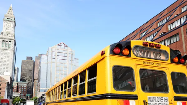 A yellow school bus drives along a road with tall skyscrapers ahead.