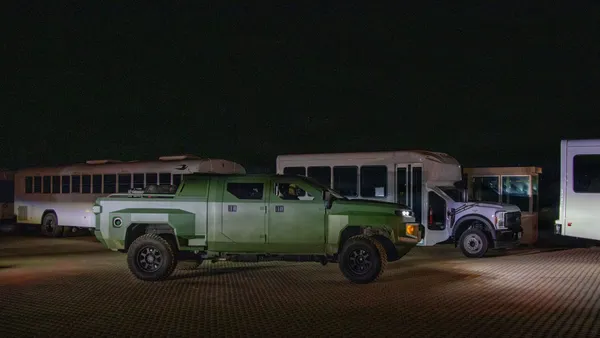 A jungle green military truck in the middle at night, with three white buses in the background.