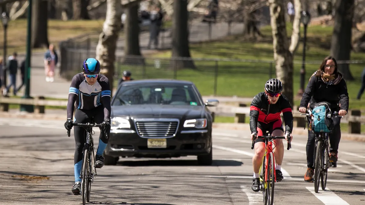 Bicyclists ride along a road with a car behind them.