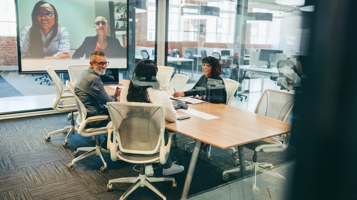 A group of employees sit in a conference room, talking with coworkers on a virtual call.