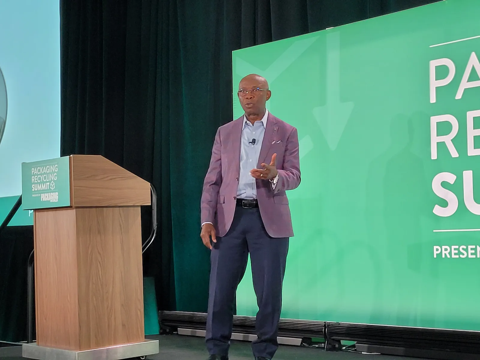 A person on a stage addresses an audience while standing in front of a green background that says Packaging Recycling Summit.