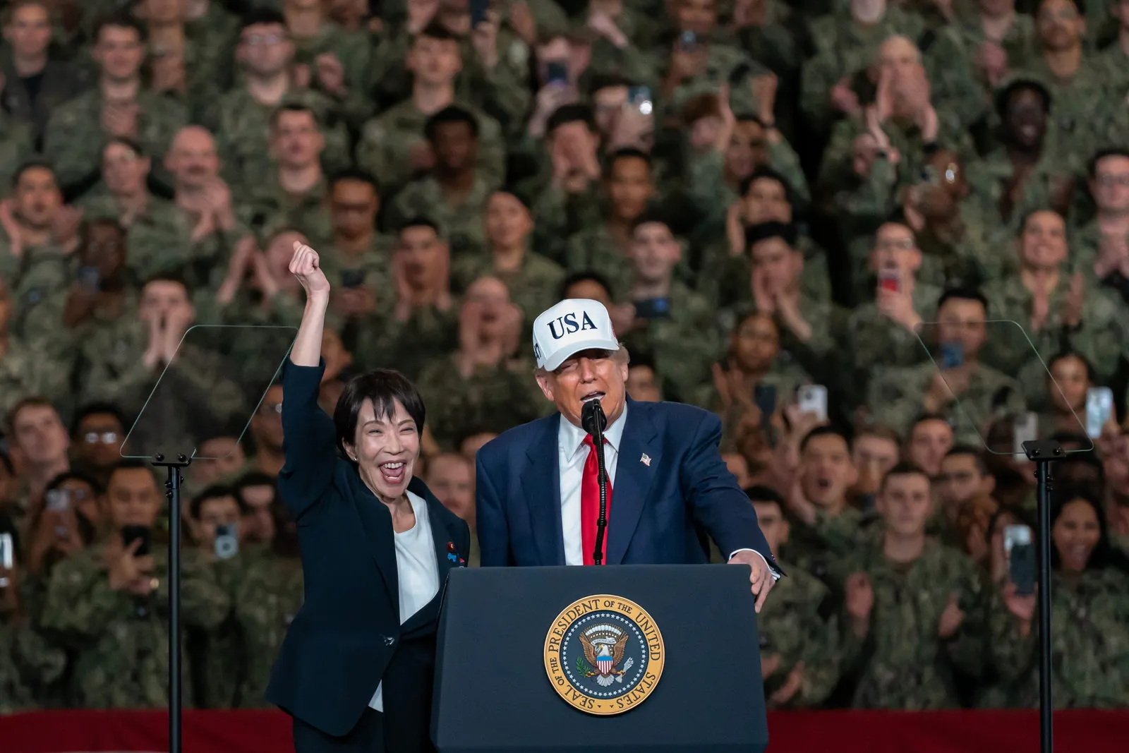Two people stand beside a pulpit, one has their hand raised and the other wears a hat saying “USA.” Dozens of people wearing green stand in the background.