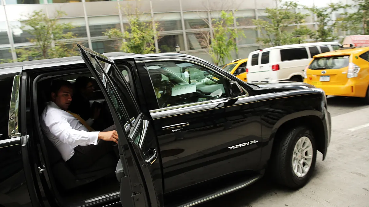 A man seated in the rear of a black car holding the door open on a busy street with yellow taxis and other vehicles.